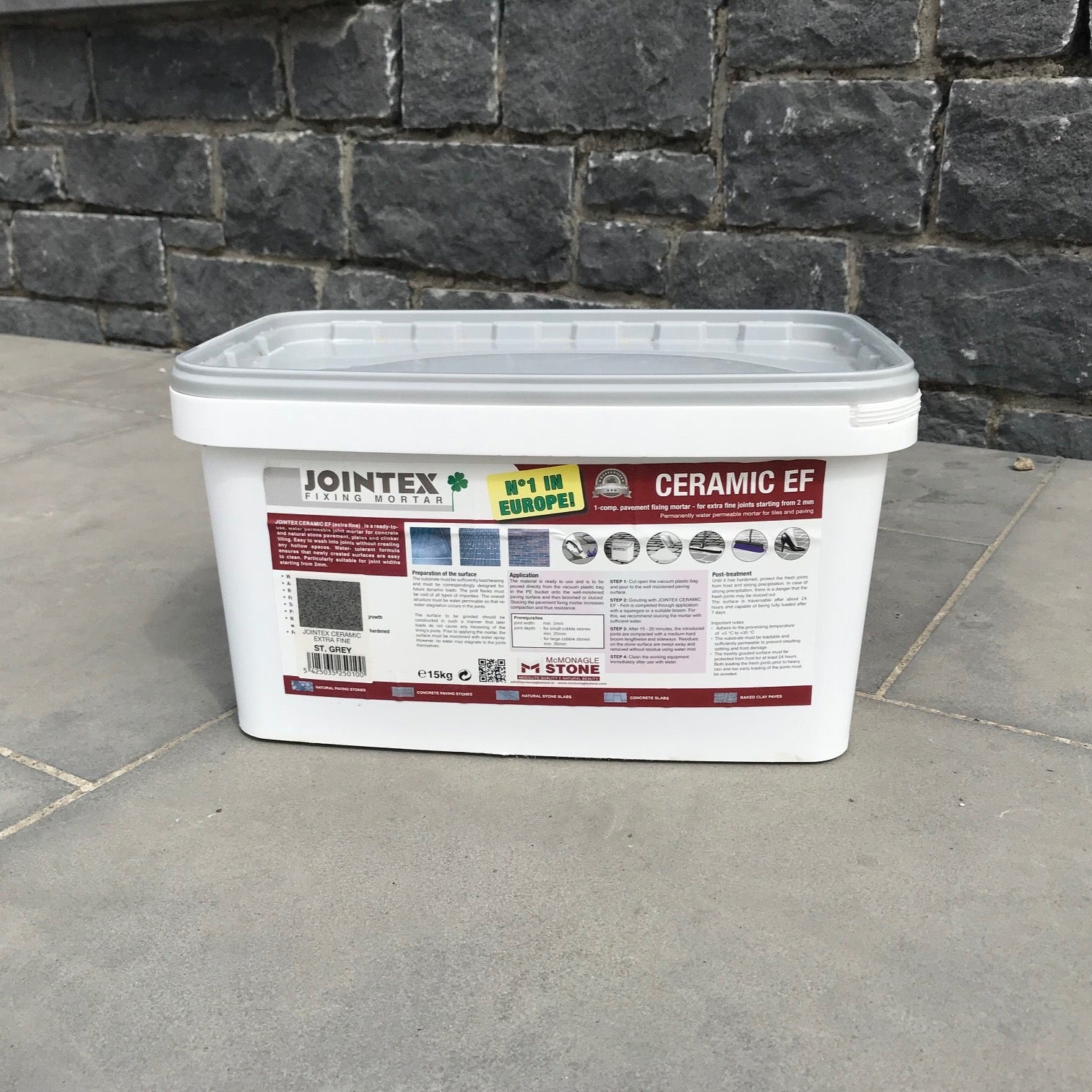 JOINTEX Easy Extra Fine Stone Grey 15kg Tub on grey paving with a dark stone wall in the background.