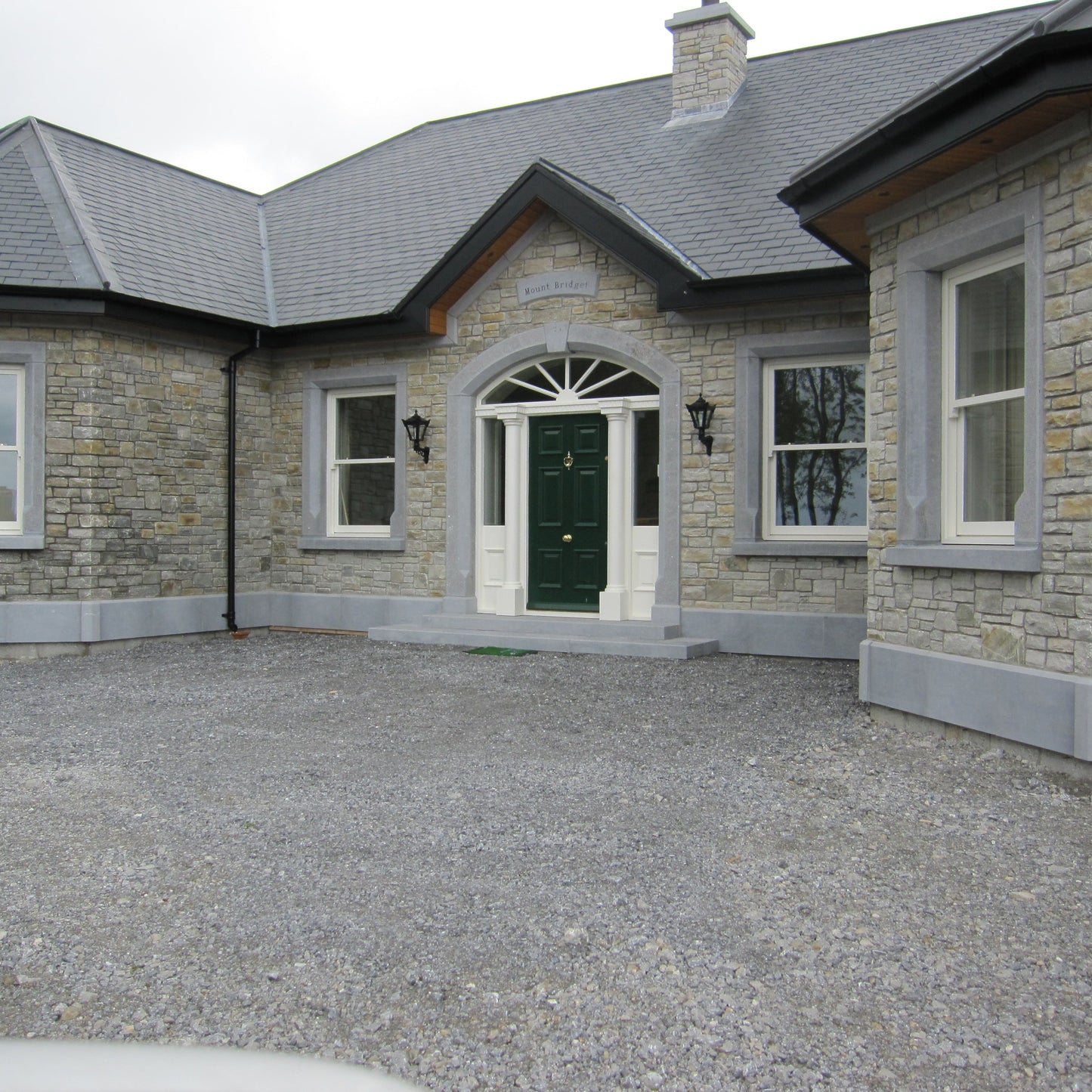 Stone house with a gravel driveway on a cloudy day. House features bespoke blue limestone door and window surrounds with a matching plinth and steps. 