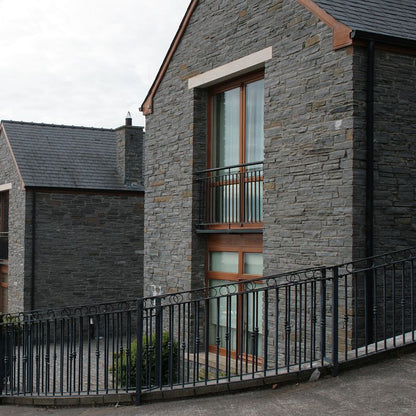 A external wall of a building constructed with Liscannor Slate, showing a gray stone finish with occasional brown variations, a window with a brown frame, and a black metal fence in the foreground.