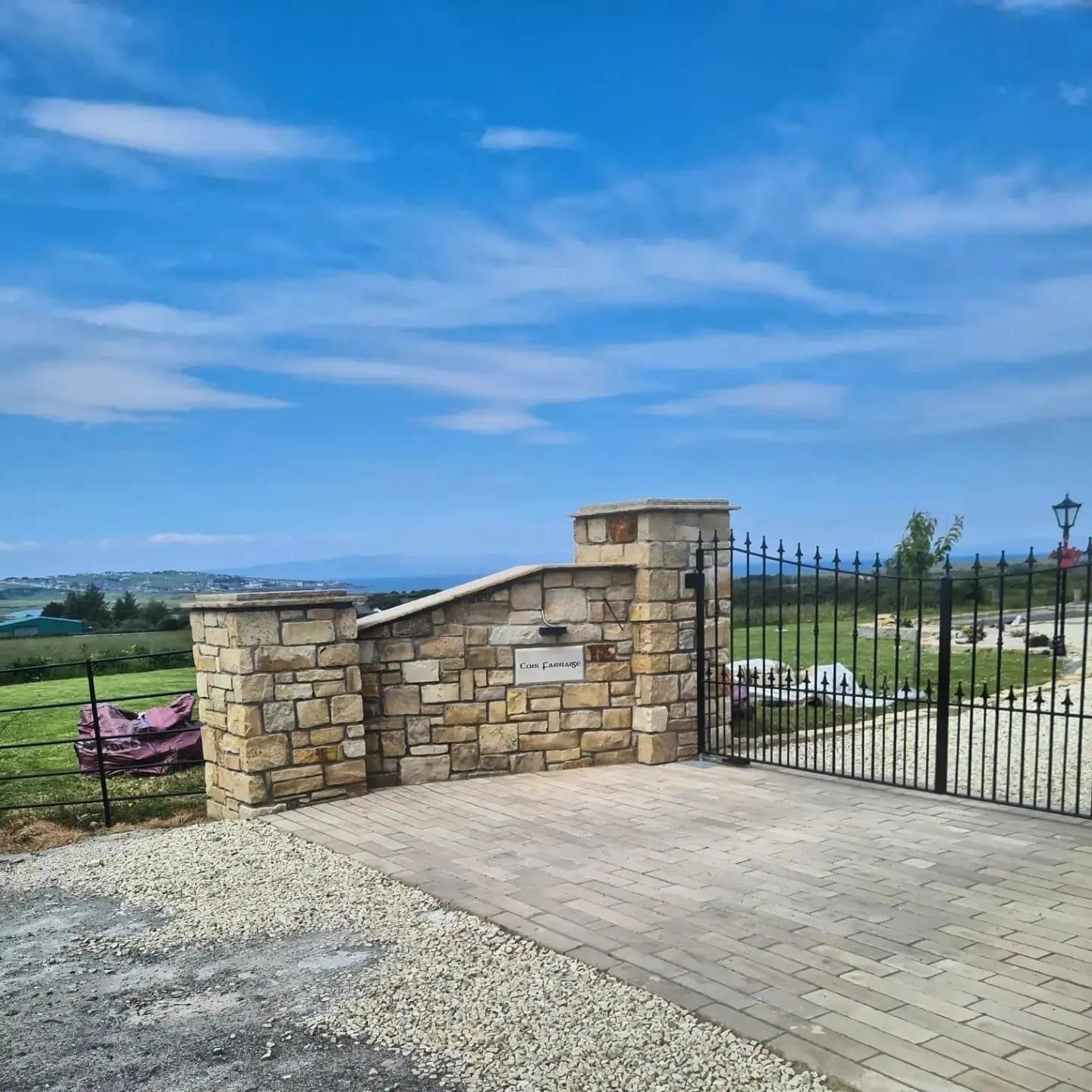 Stone wall with a gate and driveway leading to a scenic landscape under a blue sky. Stone is Mountcharles Sandstone Machined.