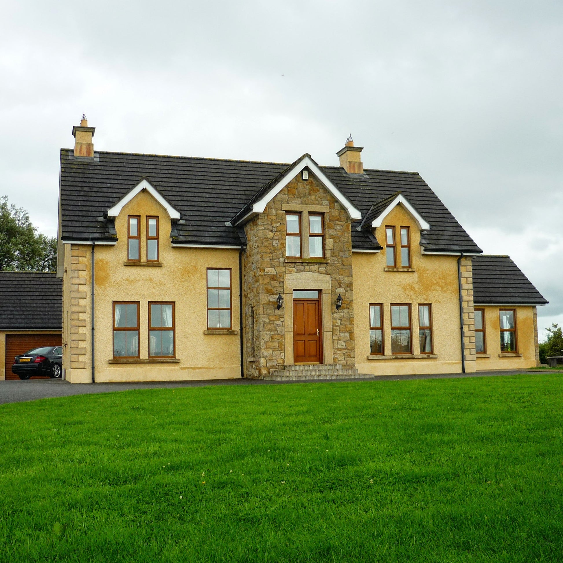 Large house with beige exterior and dark roof on a grassy lawn. Stone is Mountcharles Sandstone Machined.