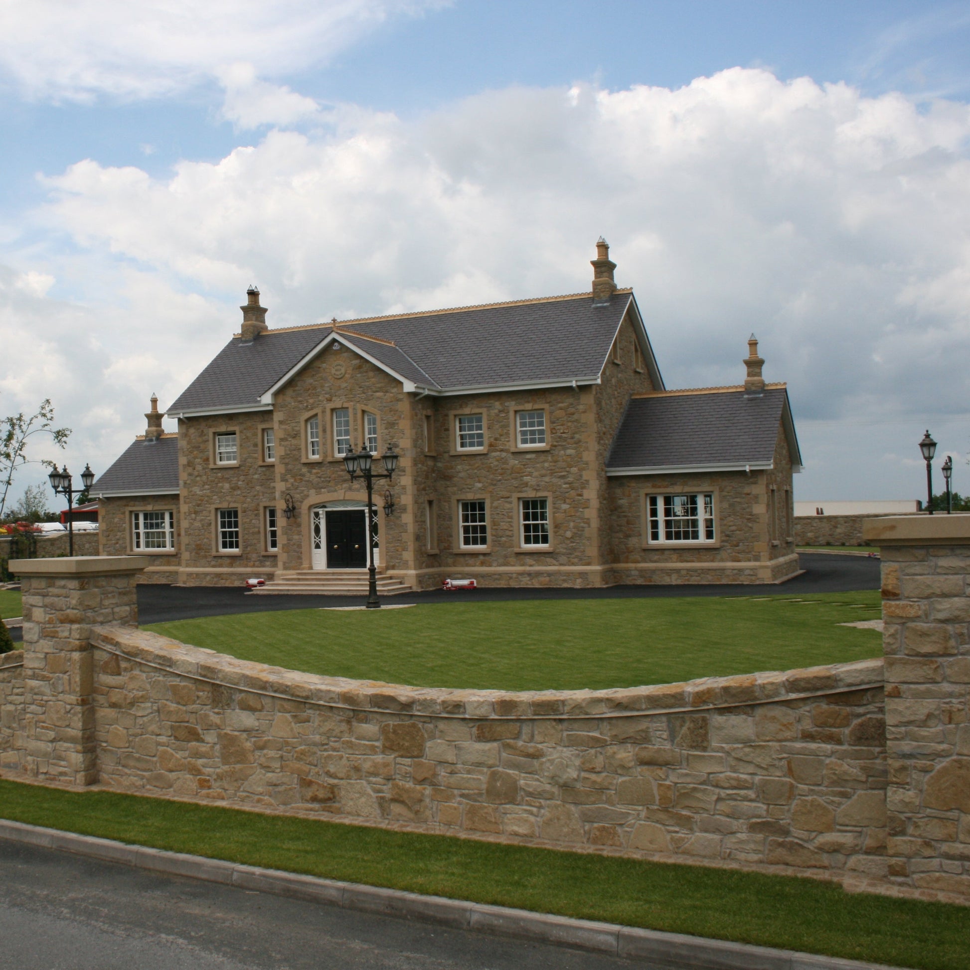 Large house with a stone wall and well-maintained garden under a blue sky with clouds. Stone is Mountcharles Sandstone Random Rubble. 