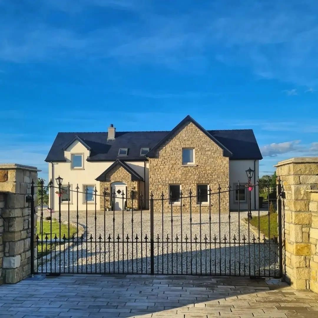 Driveway entrance with stone wall and black gate completed using Mountcharles Sandstone Paving Setts in Random Length x 100mm. Large two storey house and blue sky in the background. 