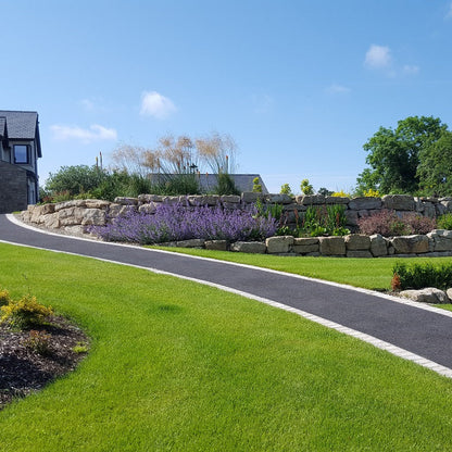 Landscape with a driveway leading to a house, surrounded by greenery and tiered Mountcharles Sandstone Rockery Stones. 