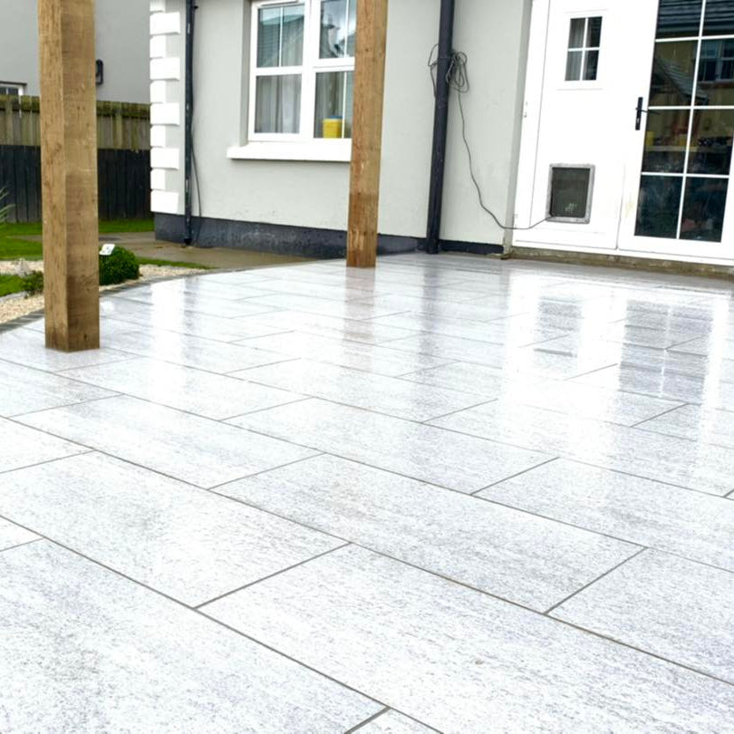 Paved patio area in a garden with a house in the background. Paving is Pietra di Bagnolo porcelain tiles. 