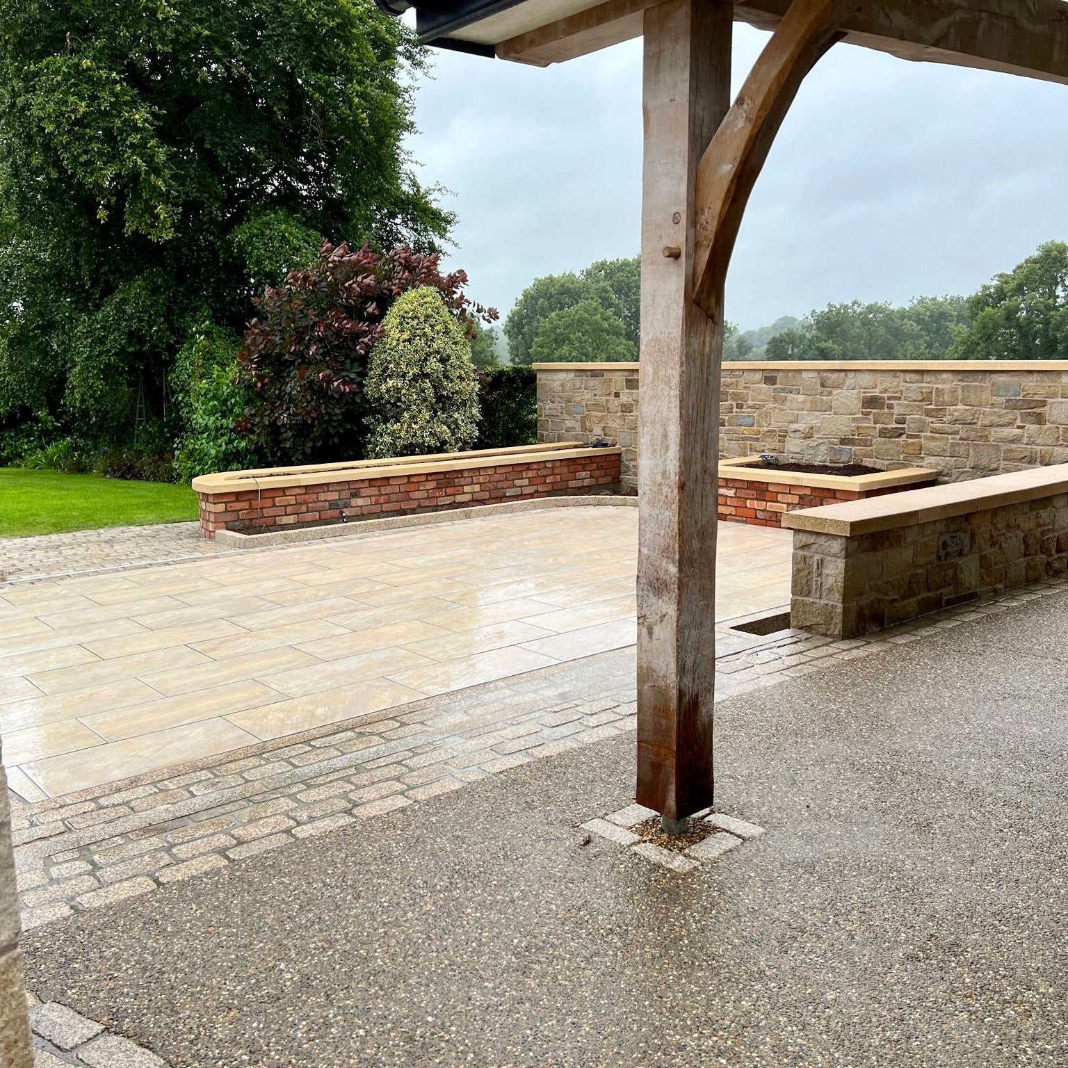 Patio area featuring Pietra Di Barge porcelain tiles and gold granite cobbles. Trees and red brick planters in the background. 