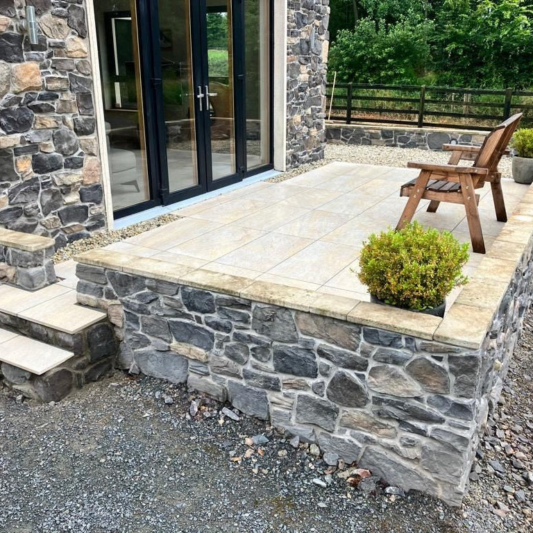 Raised patio area with steps and brown wooden bench with a potted plant on either side. Paving is Pietra Di Barge porcelain tiles. 