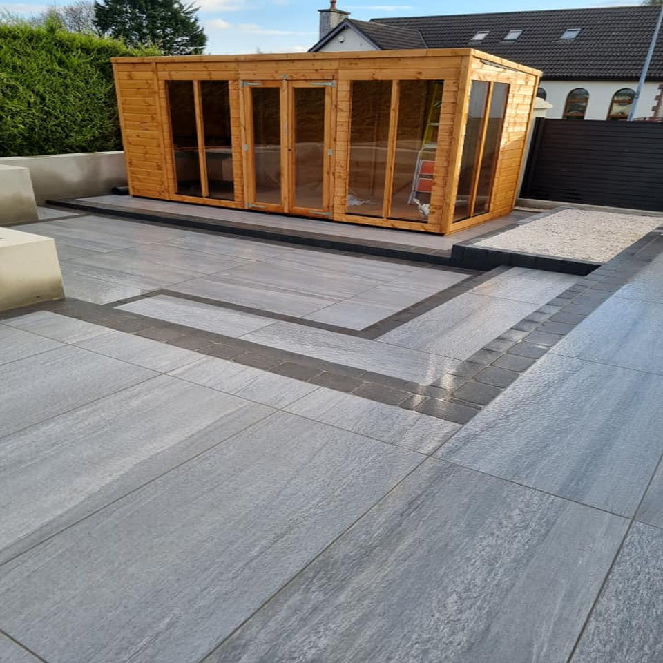 An outdoor installation of Pietra di Vals Porcelain Paving in grey, laid out on a garden patio area with a wooden shed in the background. 