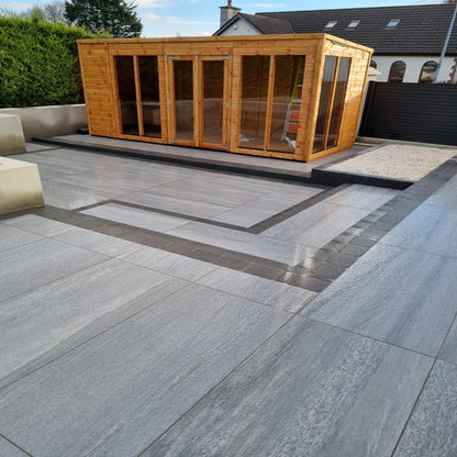 An outdoor installation of Pietra di Vals Porcelain Paving in grey, laid out on a garden patio area with a wooden shed in the background. 