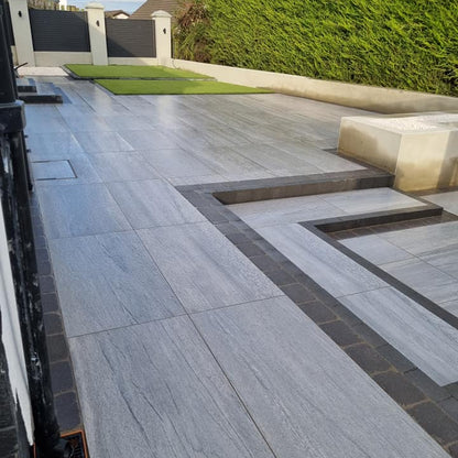 Large patio featuring Pietra Di Vals Porcelain Tiles with hedging and fence in the background.