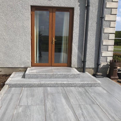 Grey stone pathway leading to a wooden door with glass panels on a grey building. Paving is Pietra di Vals porcelain tiles.