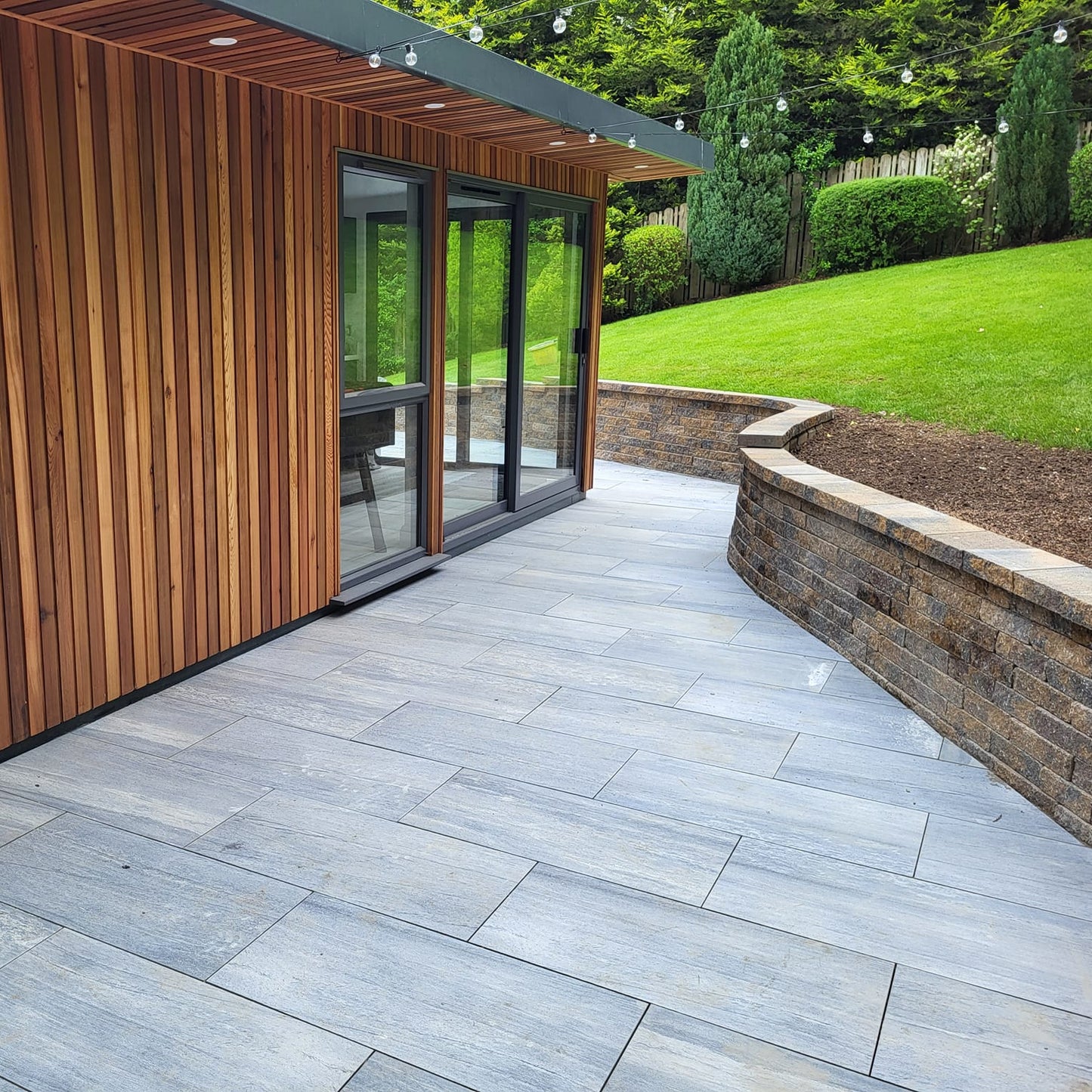 Patio area with stone wall and wooden cladded house. Paving is Pietra di Vals porcelain tiles.