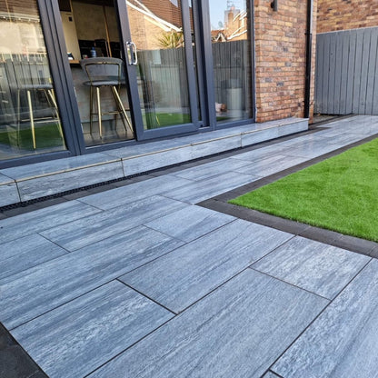 An outdoor installation of Pietra di Vals porcelain tiles in a garden setting, showcasing their dark grey colour and rectangular shape.
