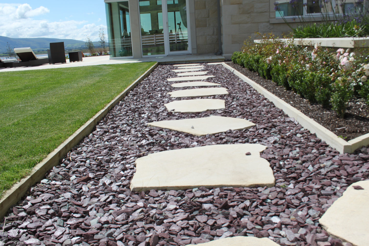 Garden pathway with stepping stones leading to a stone house with large windows. Pathway features Plum Slate Mulch. 