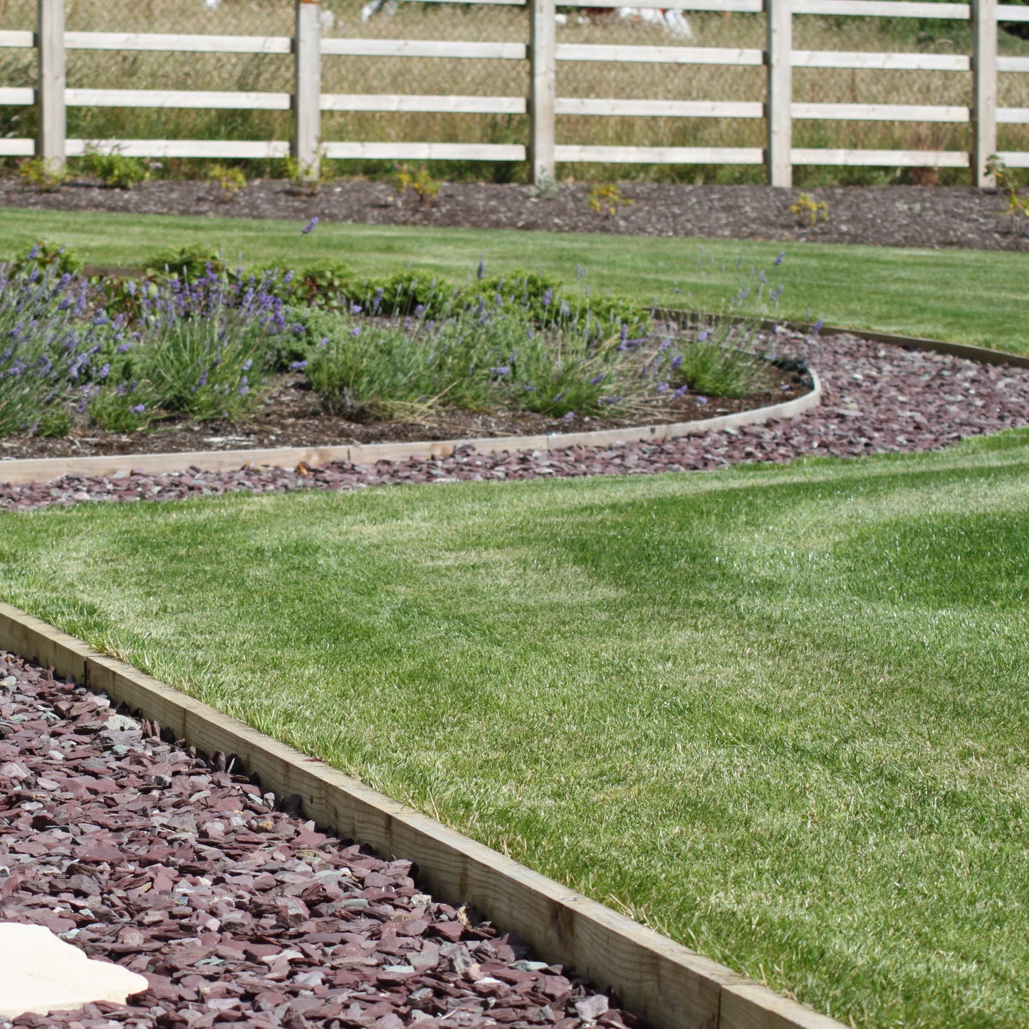 Garden with a curved stone path, grass, and a white fence in the background. Pathway and garden beds feature plum slate mulch decorative garden stone. 