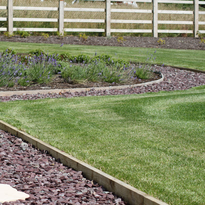 Garden with a curved stone path, grass, and a white fence in the background. Pathway and garden beds feature plum slate mulch decorative garden stone. 