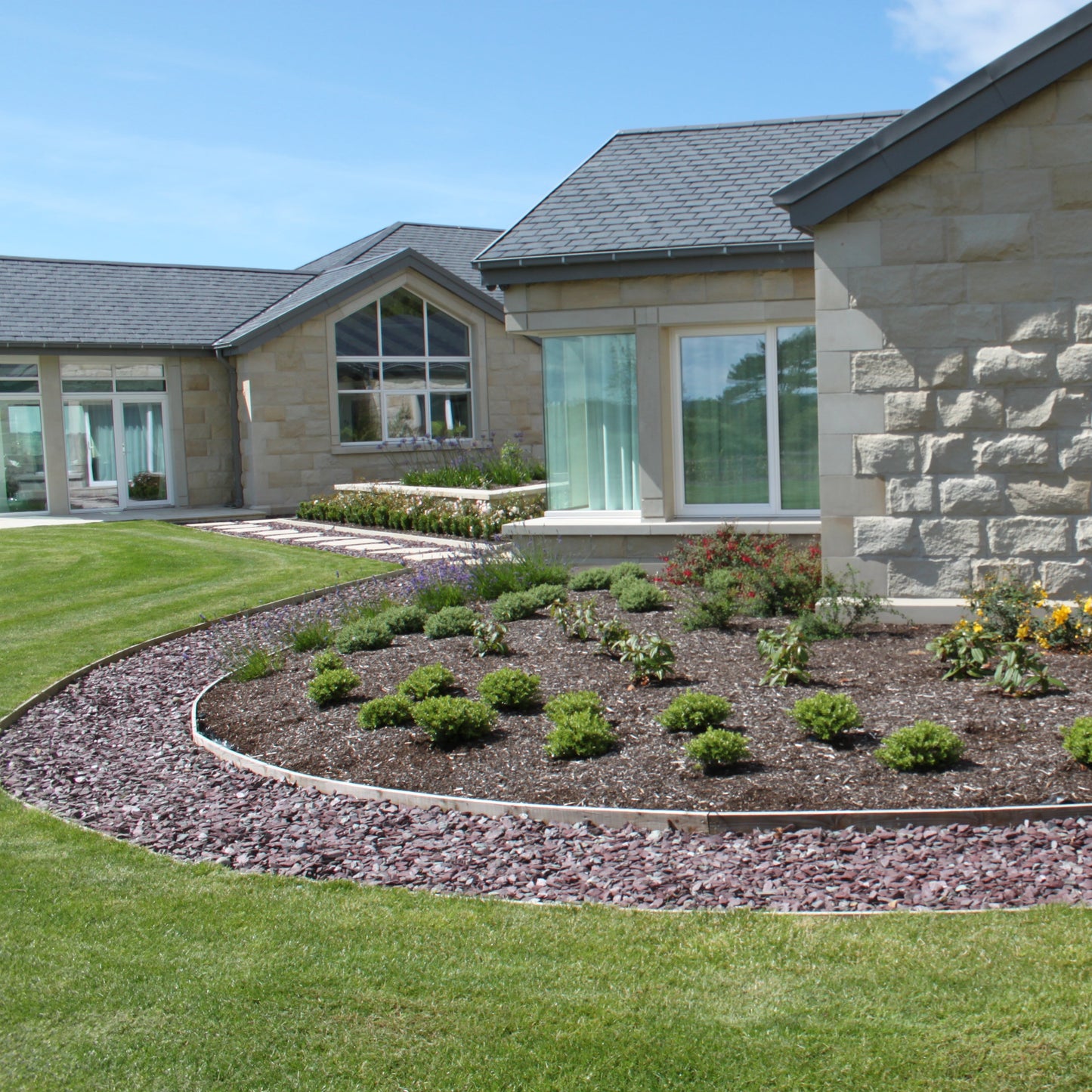 Neatly landscaped garden in front of a house with a clear blue sky. Pathway features Plum Slate Mulch. 