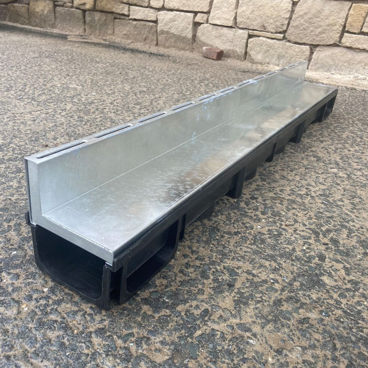 Galvanised Poly Brick Slot Channel Drain on stone pavement with a natural stone wall in the background.