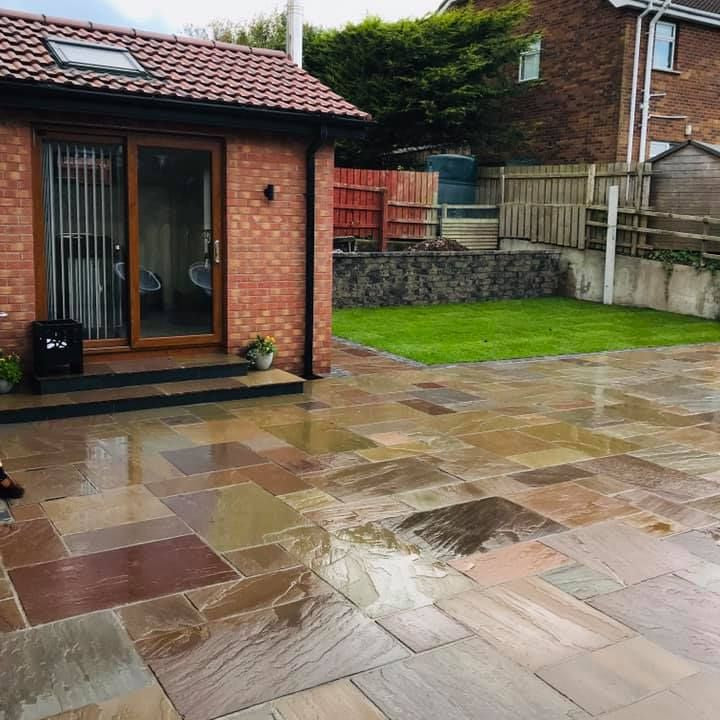 Paved patio area featuring Raj Green Sandstone paving in a garden with a brick house and green lawn in the background.