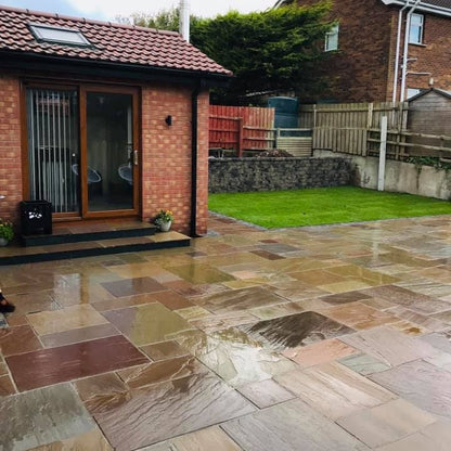 Paved patio area featuring Raj Green Sandstone paving in a garden with a brick house and green lawn in the background.