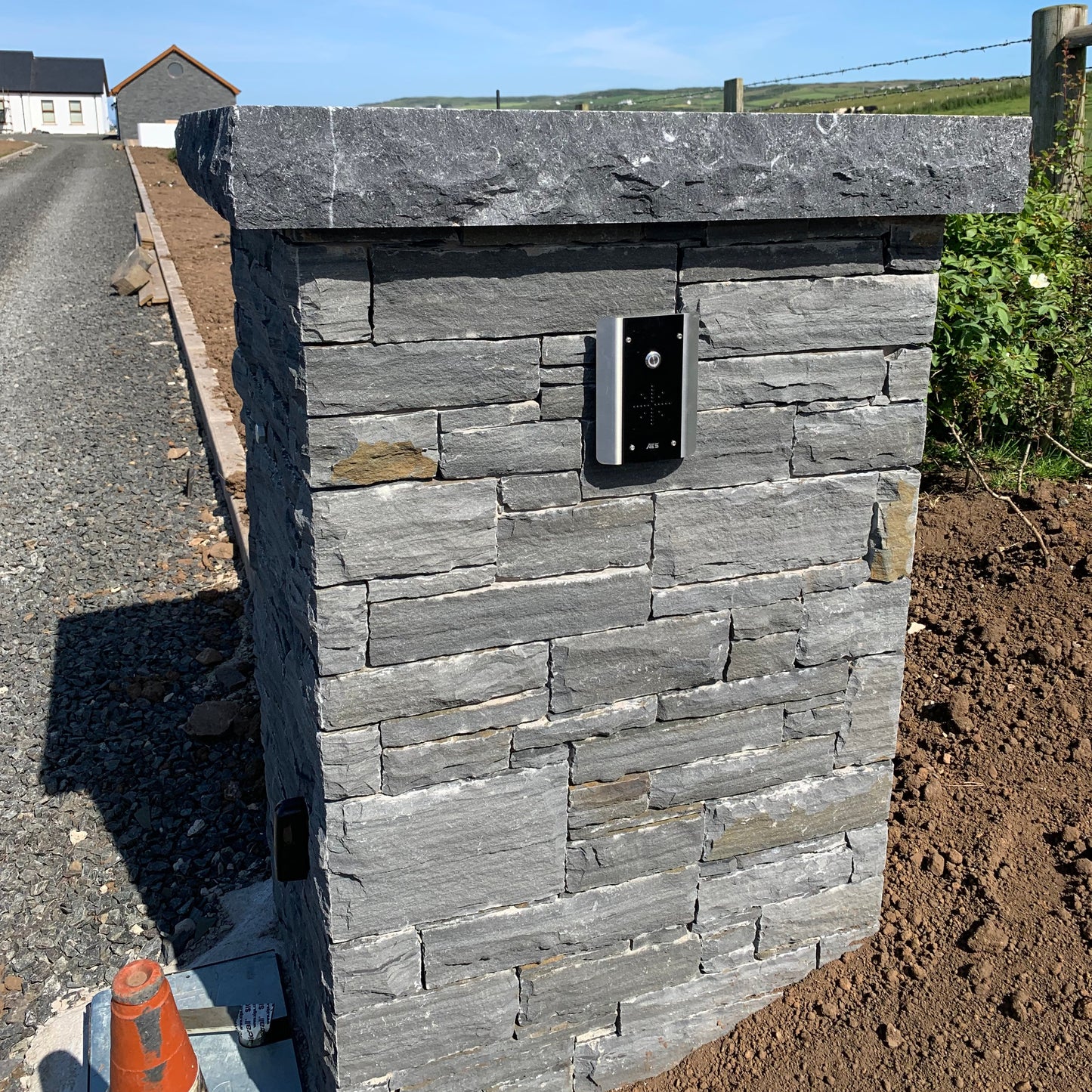 Stone wall with a small electronic device mounted on it, set against a clear blue sky. Wall completed with flat blue limestone pillar cap with rockfaced edge. 