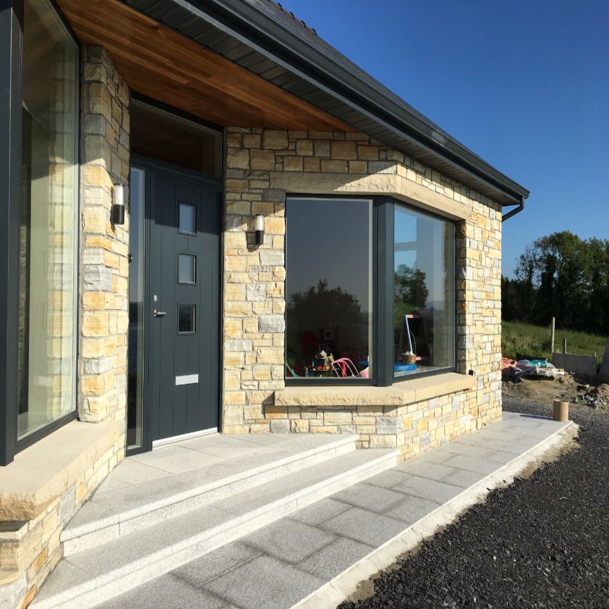 Modern house exterior with stone façade, glass windows, and a clear blue sky. Bespoke rockfaced sandstone window lintel and sills.