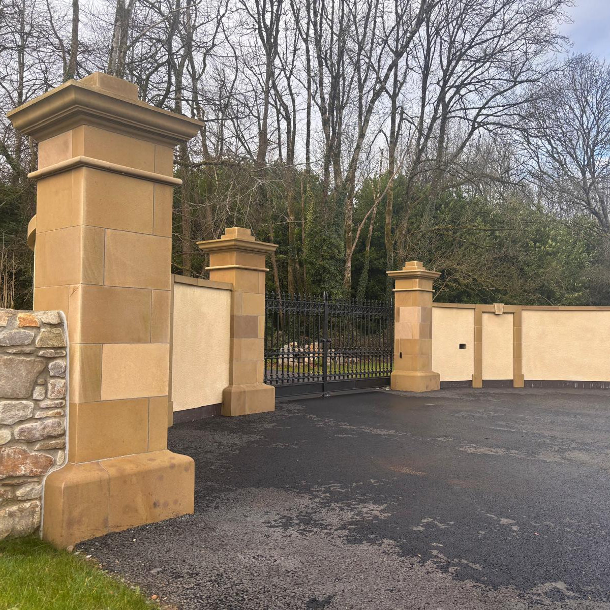 Decorative stone gate with bespoke sandstone pillars featuring plinths in a driveway, surrounded by trees and a clear sky.