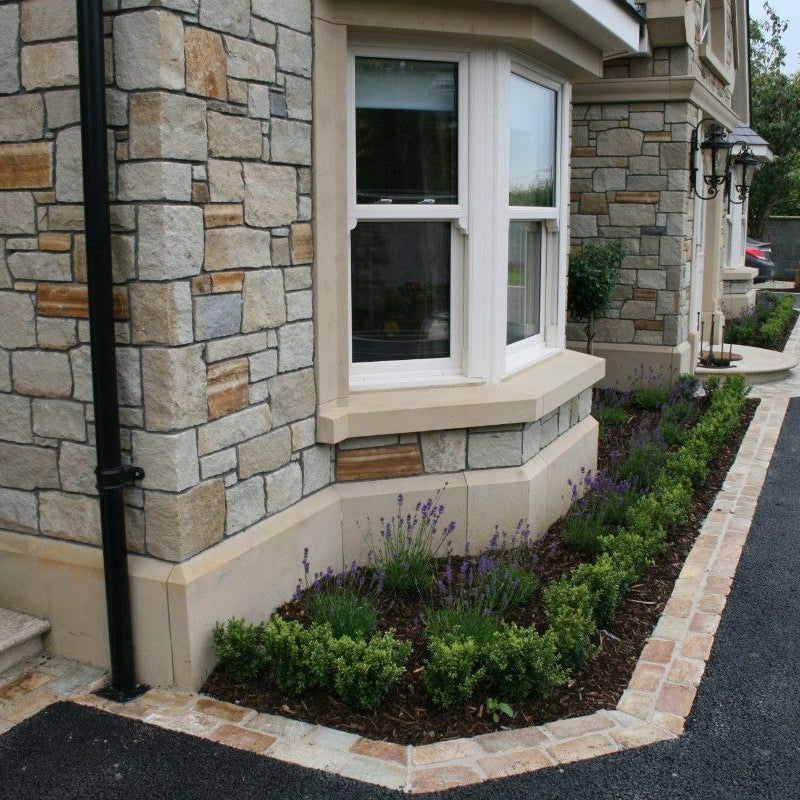 The image shows the lower part of a stone-fronted building with a classic architectural design, featuring a bespoke sandstone plinth with a planted area and a window. 