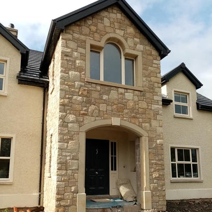 Stone house exterior with a partially completed front porch. Unique sandstone window surround above door surround. 