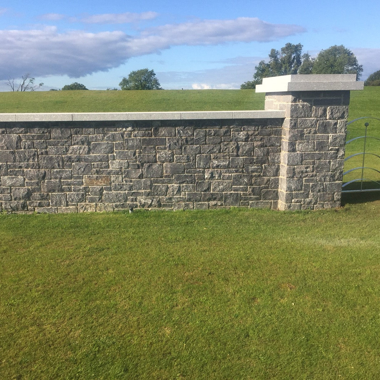 Stone wall with a metal gate in a grassy field under a blue sky with clouds. Stone wall completed with Silver Granite Wall Capping.