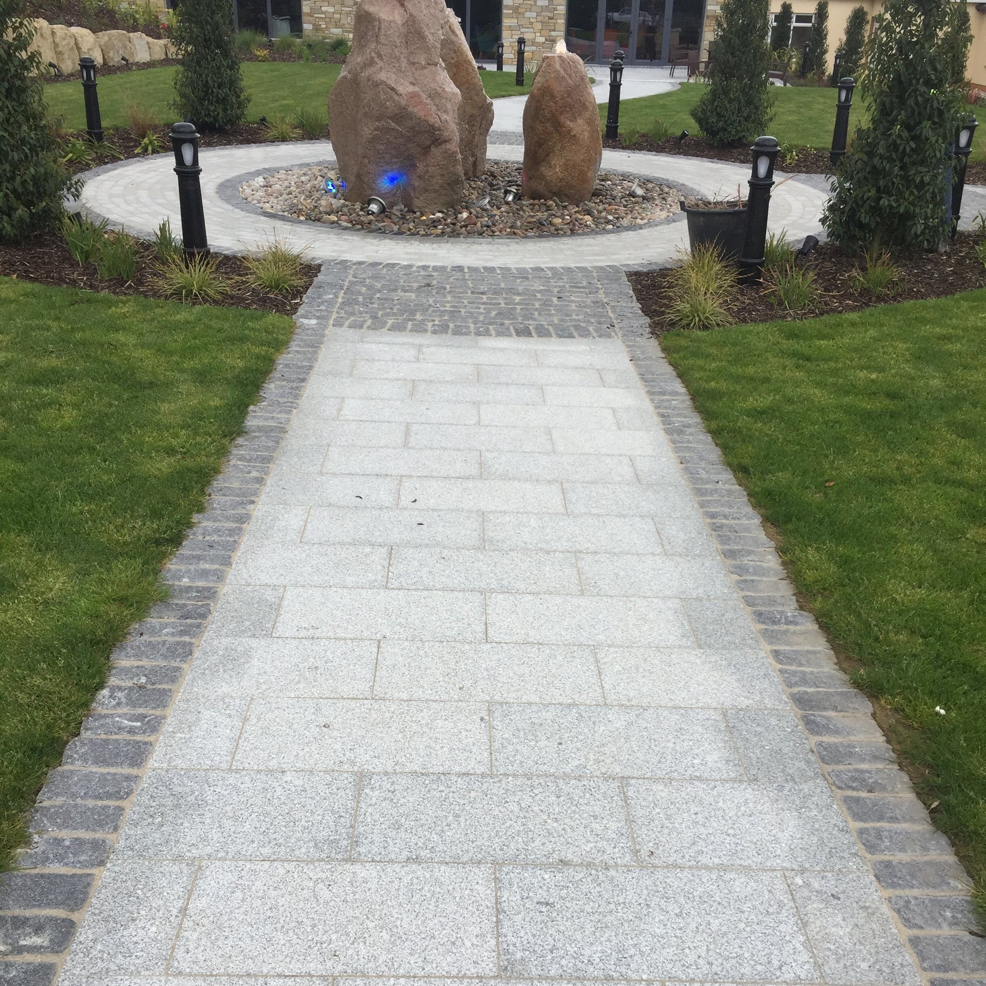 Pathway in Silver Granite Paving in 600 x 300mm size bordered by Black Granite Cobbles in 100x100mm size leading towards a circular garden feature of Sandstone Monoliths. 
