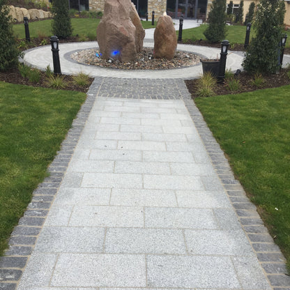 Pathway in Silver Granite Paving in 600 x 300mm size bordered by Black Granite Cobbles in 100x100mm size leading towards a circular garden feature of Sandstone Monoliths. 