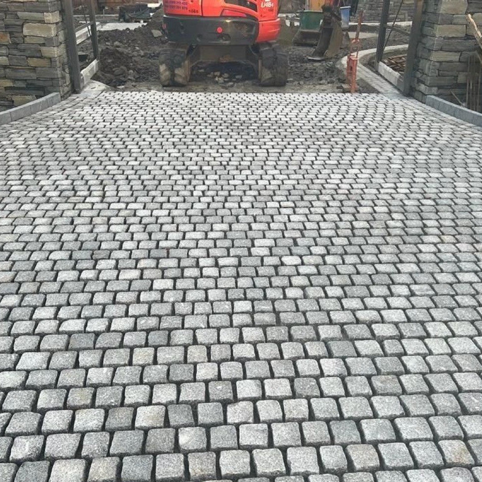 Paved area being installed using Silver Granite Paving Cobbles with a digger in the background between two stone pillars. 