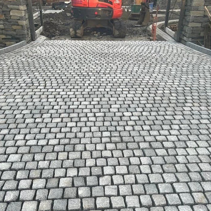 Paved area being installed using Silver Granite Paving Cobbles with a digger in the background between two stone pillars. 