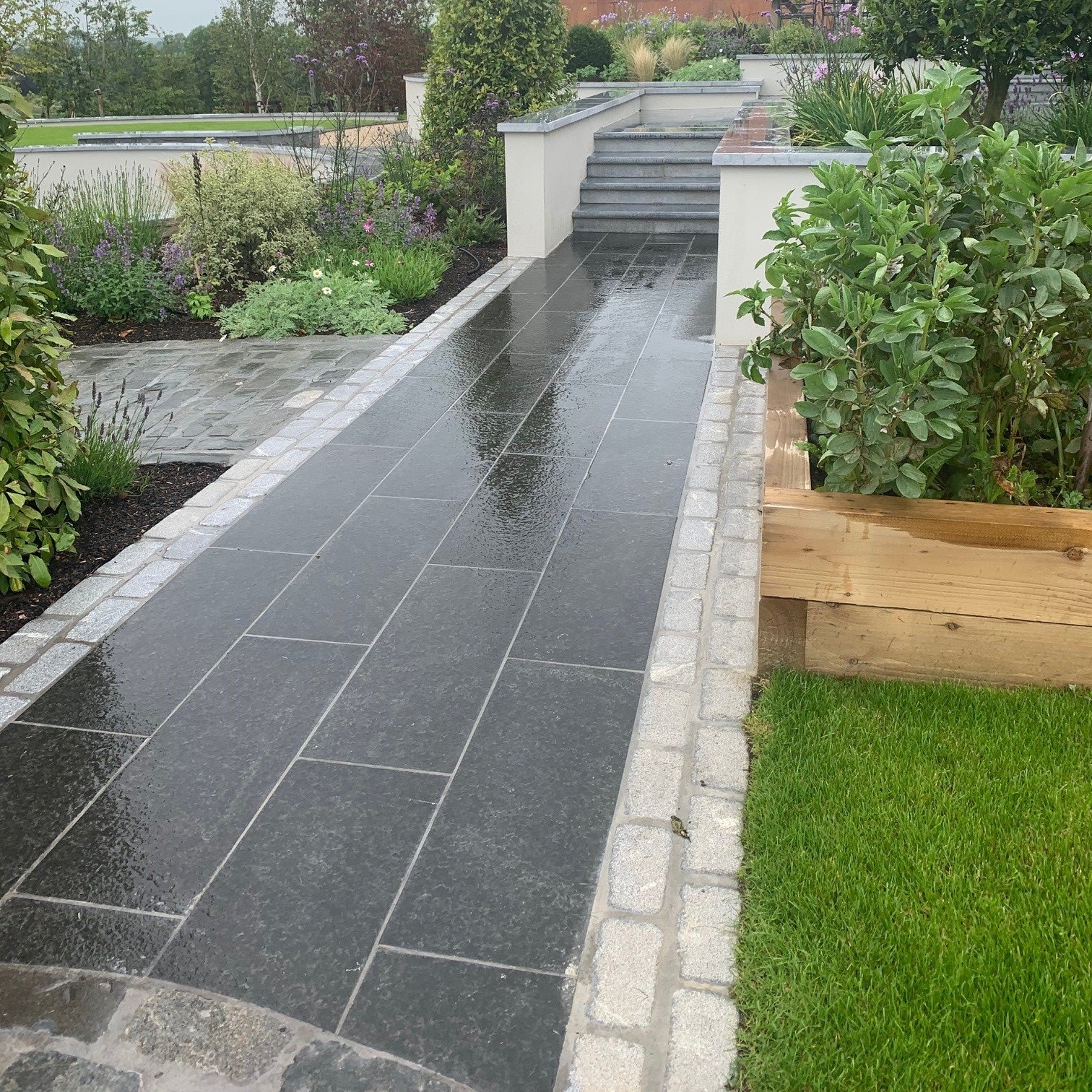 Garden pathway completed using dark basalt paving and silver granite cobbles on either side. Lots of planting and trees in the background. 