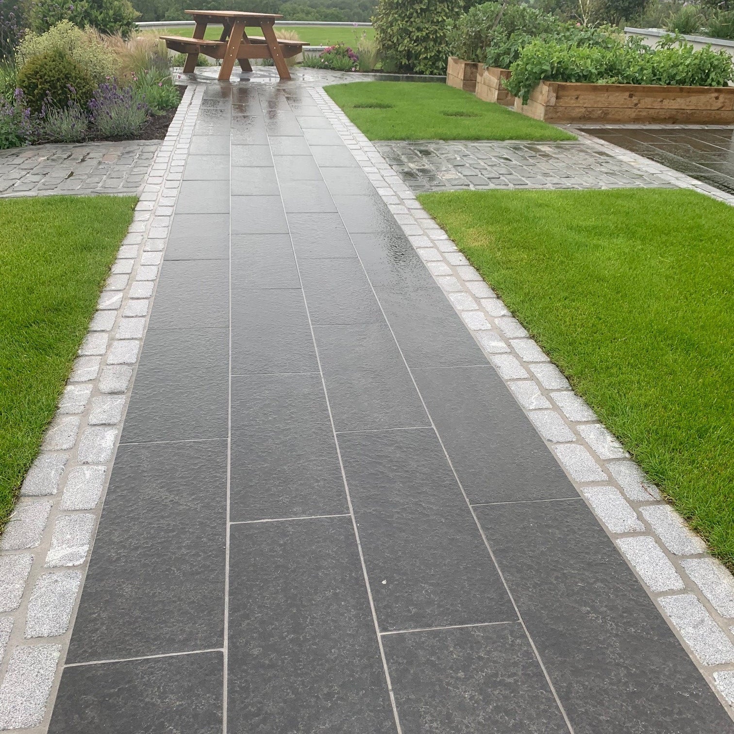 Paved pathway with grass on either side leading towards a picnic bench. Pathway edging completed using Silver Granite Cobbles in 200 x 100mm  