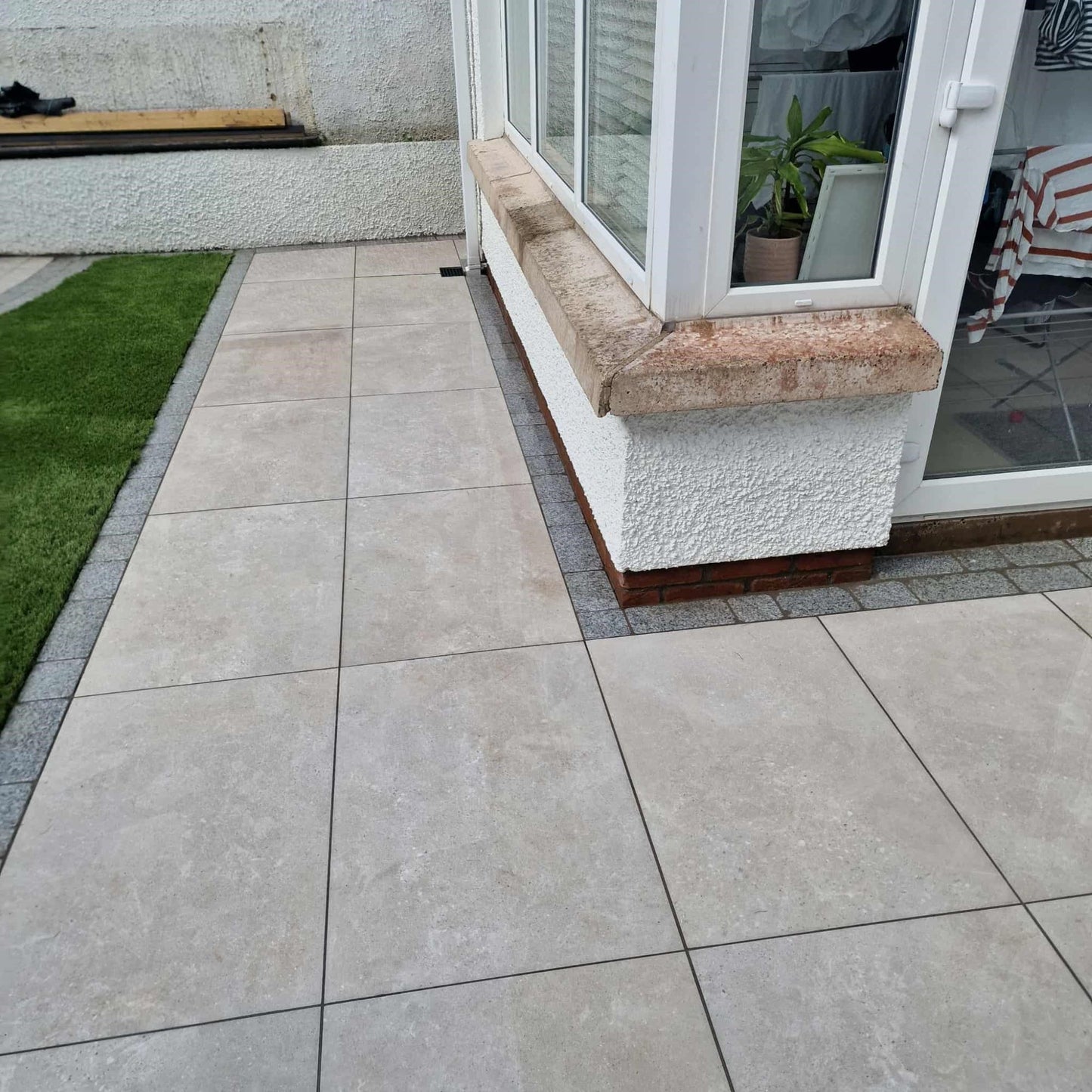 Tiled patio area featuring Silver Street Range Porcelain Paving tiles with a grassy patch and a house entrance.