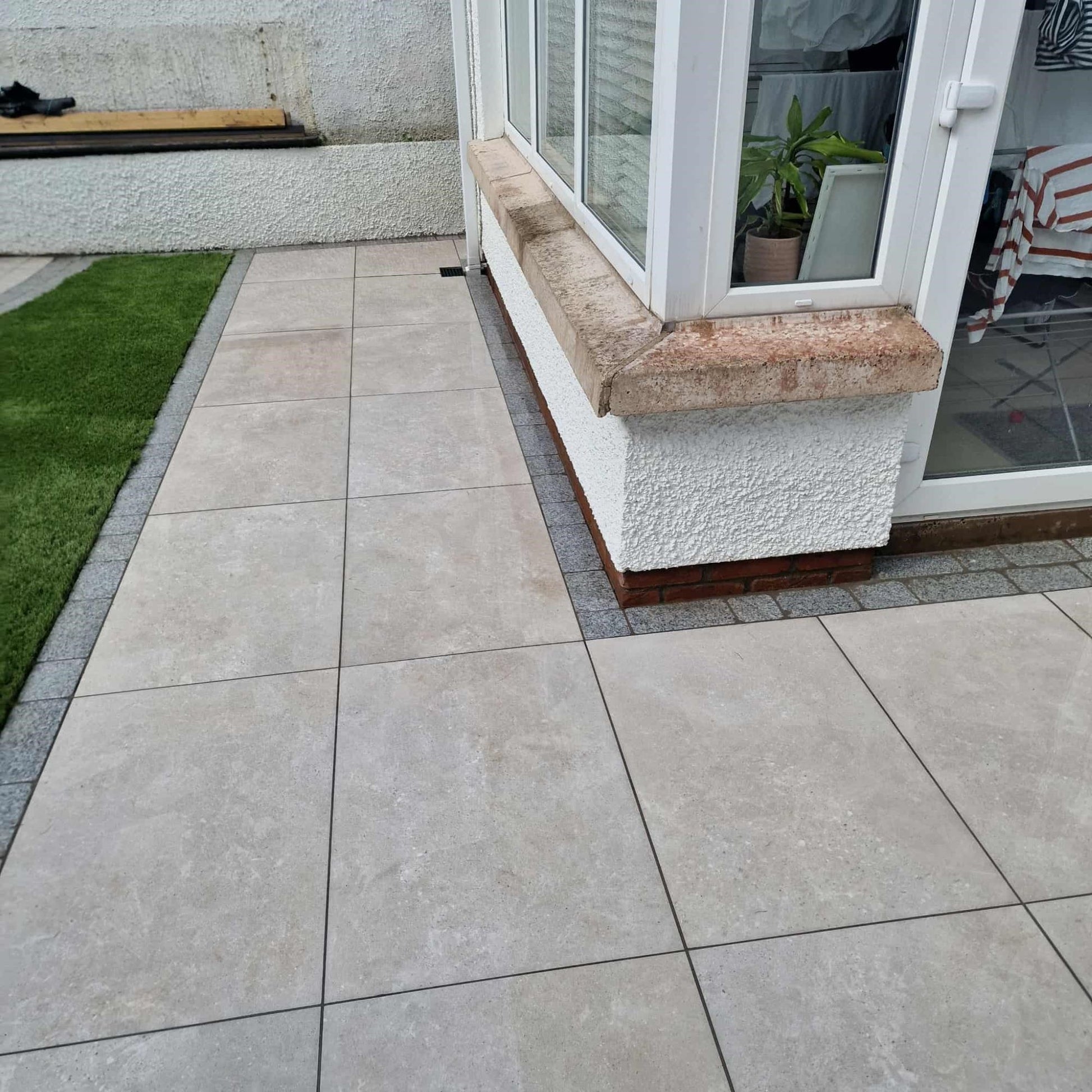 Tiled patio area featuring Silver Street Range Porcelain Paving tiles with a grassy patch and a house entrance.