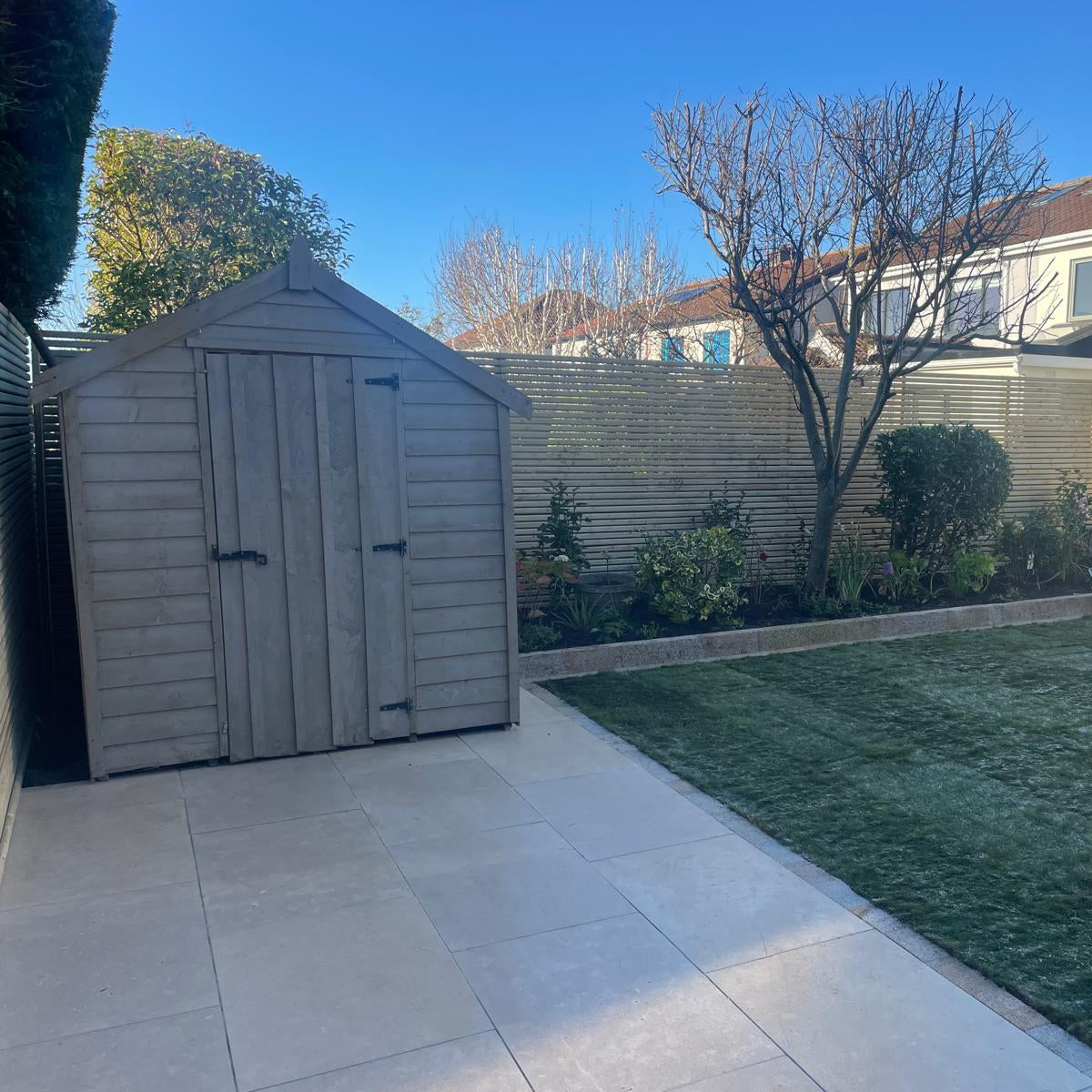 Backyard with a wooden shed, paved patio, and grass area on a clear day. Paving is Sinai Pearl Porcelain Paving tiles. 
