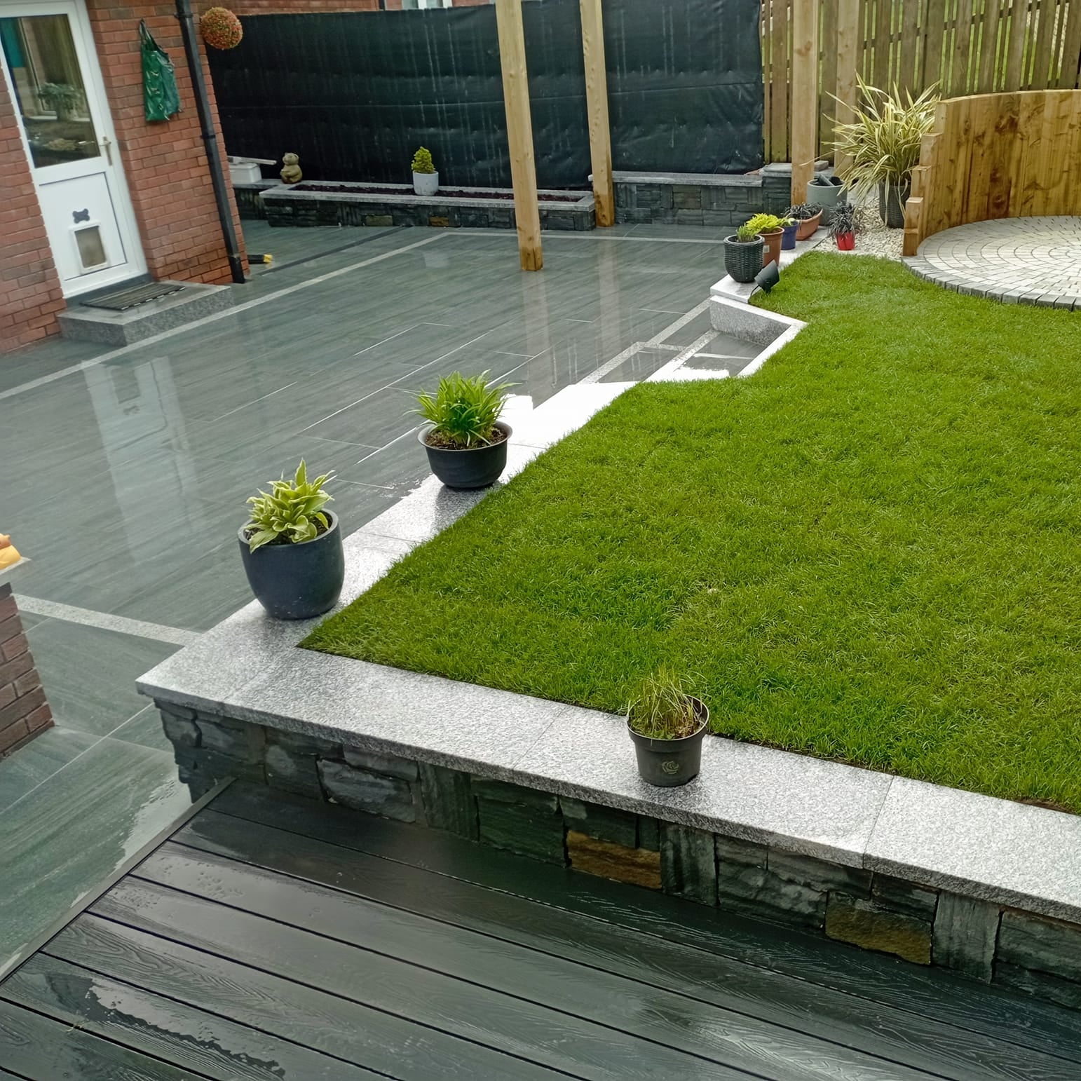 Patio area and raised garden with lots of potted plants dotted around. Paving is Smokey Jewel Range Porcelain Paving. 