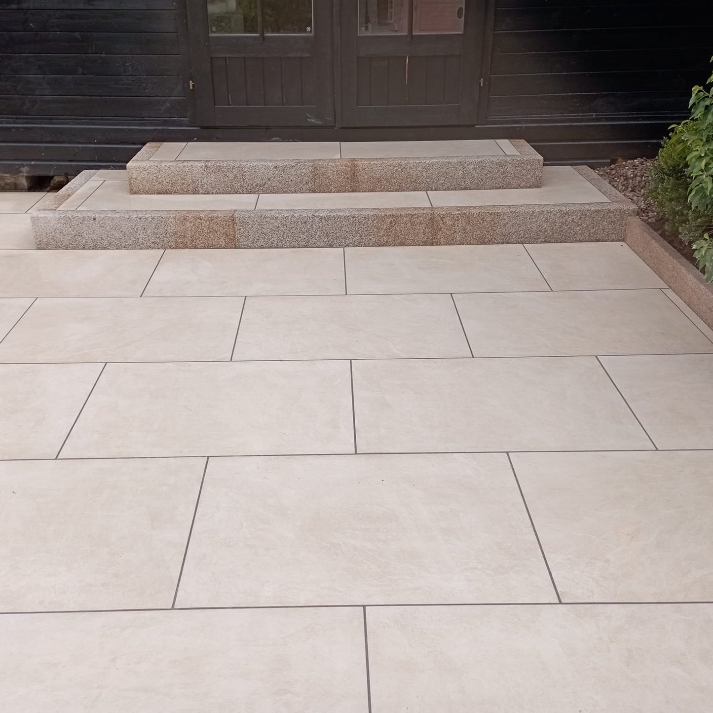 Patio with steps leading up to a black wooden shed. Paving is Station White Jewel Range Porcelain Paving Tiles. 