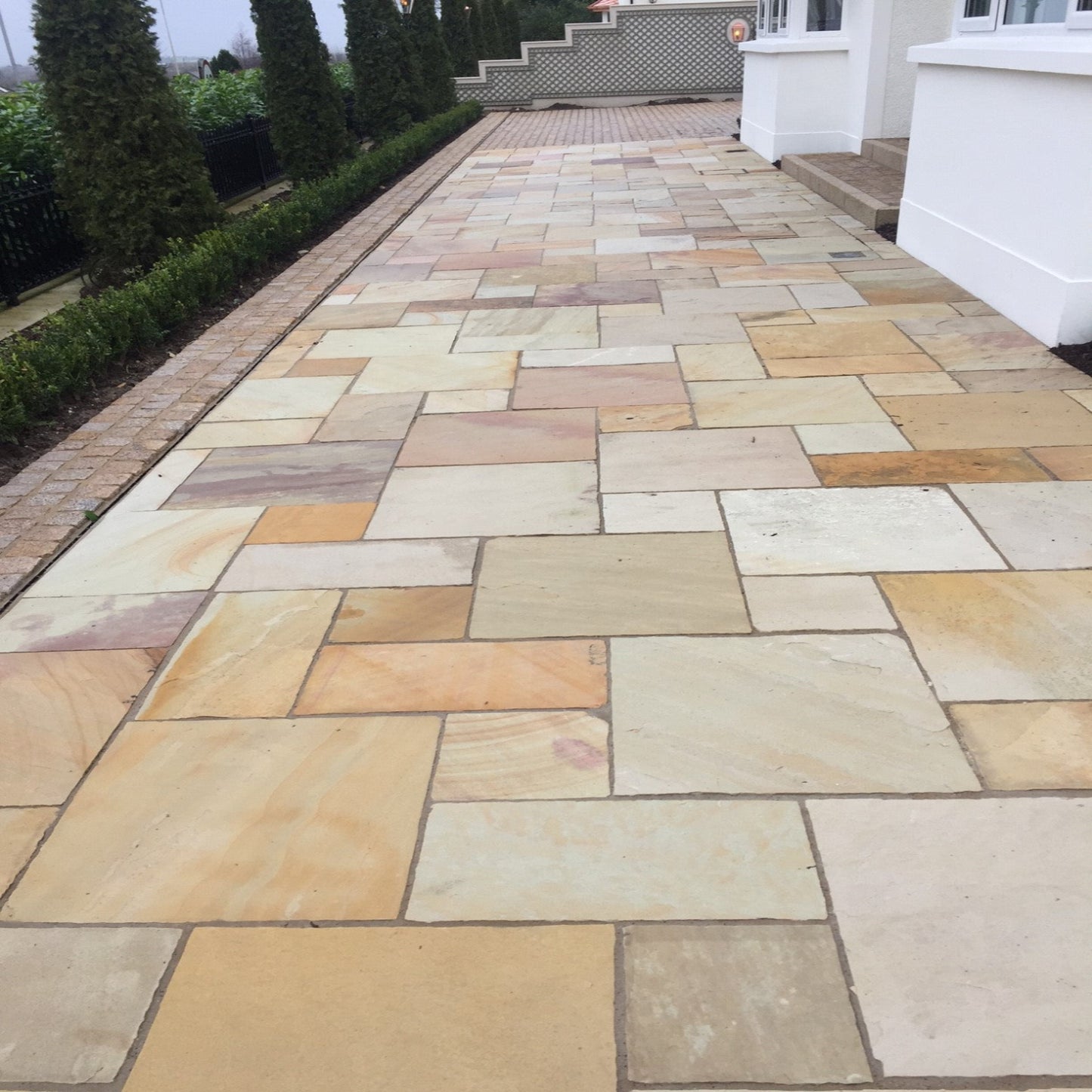 A Tinted Mint Sandstone Patio Pack installed on a garden pathway, showcasing a natural stone with varying shades of yellow, buff, pink, and golden brown tones.