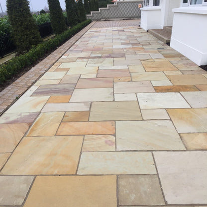 A Tinted Mint Sandstone Patio Pack installed on a garden pathway, showcasing a natural stone with varying shades of yellow, buff, pink, and golden brown tones.