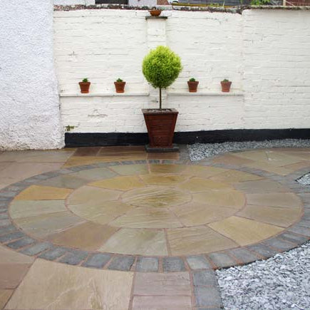 Round stone patio completed using Tinted Mint Sandstone Paving Circle with a white brick wall and potted plants in the background. 