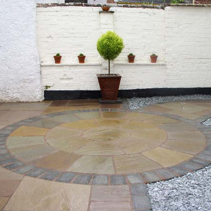 Round stone patio completed using Tinted Mint Sandstone Paving Circle with a white brick wall and potted plants in the background. 