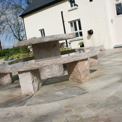 Stone outdoor table and bench set on a patio featuring 4m Tinted Mint Sandstone Paving Circle in front of a house.