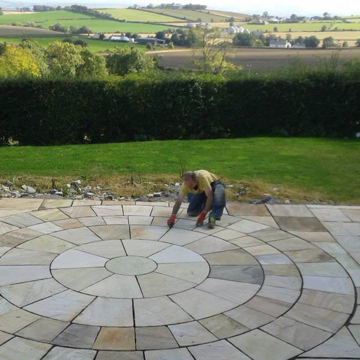 Person working on a 4m Tinted Mint Sandstone Paving Circle on a patio in a garden with a scenic background. 