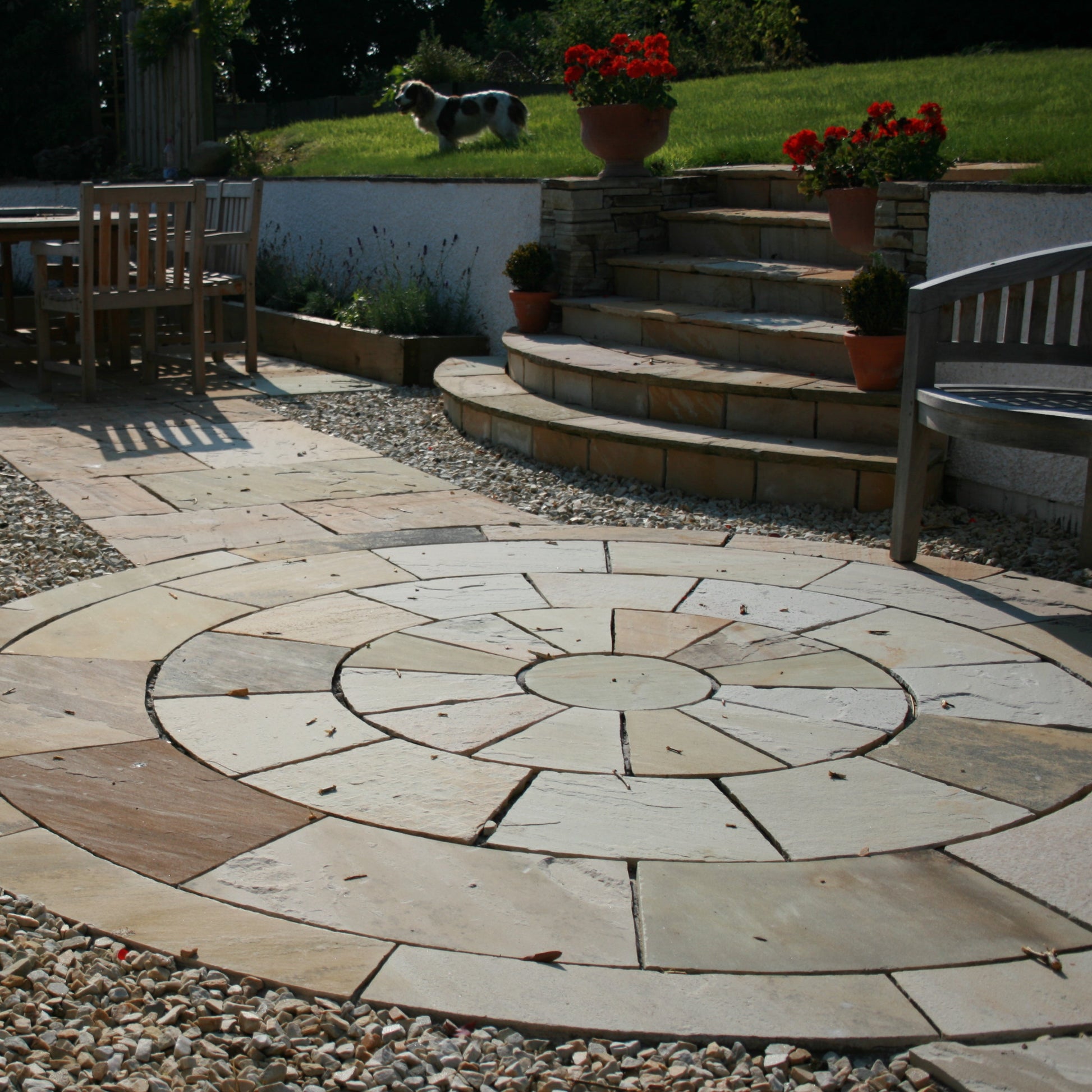 Patio area featuring a 4m Tinted Mint Sandstone Paving Circle surrounded by natural stone chipping, potted plants and garden furniture. 