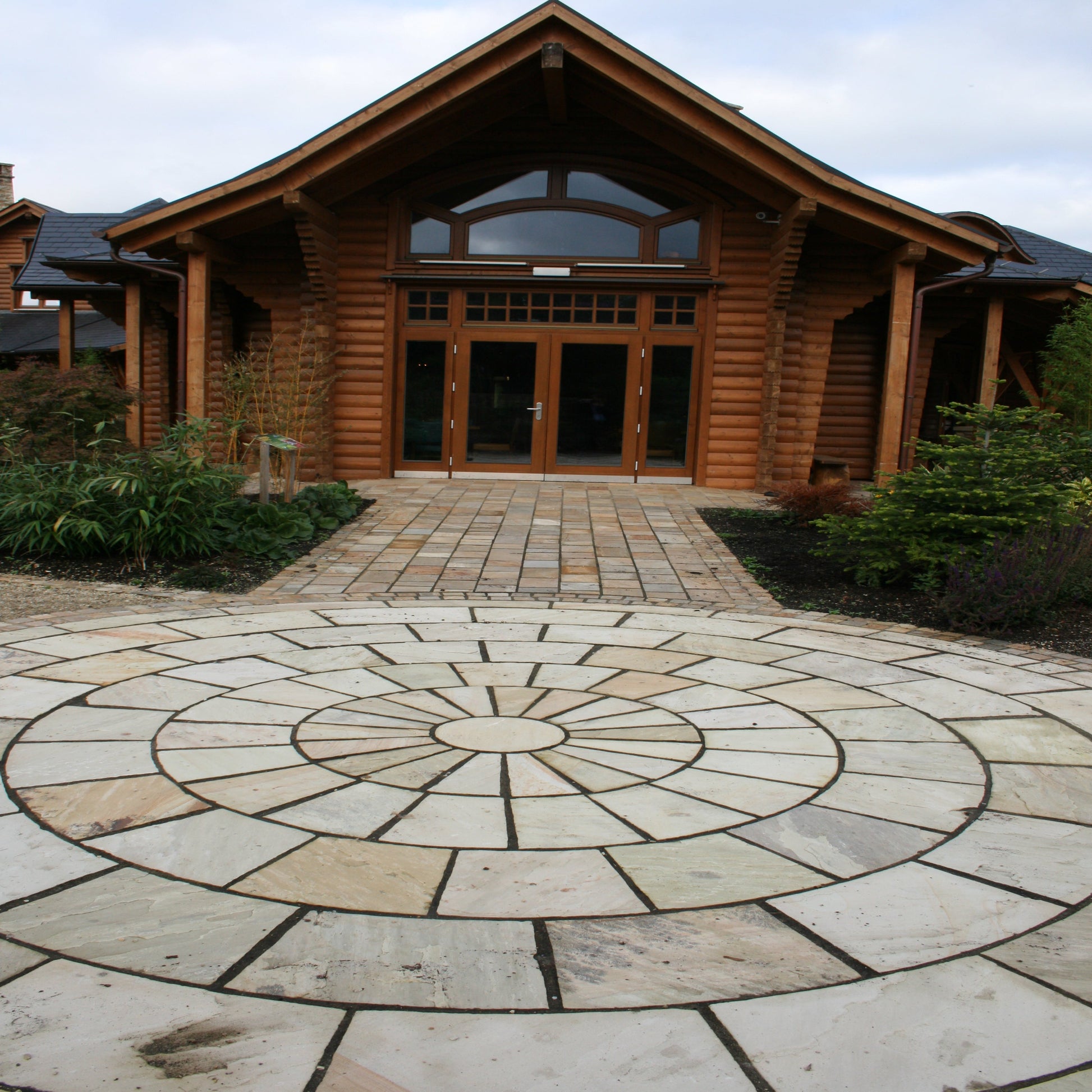 A 5m Tinted Mint Sandstone Paving Circle with pathway leading to large wooden building. 