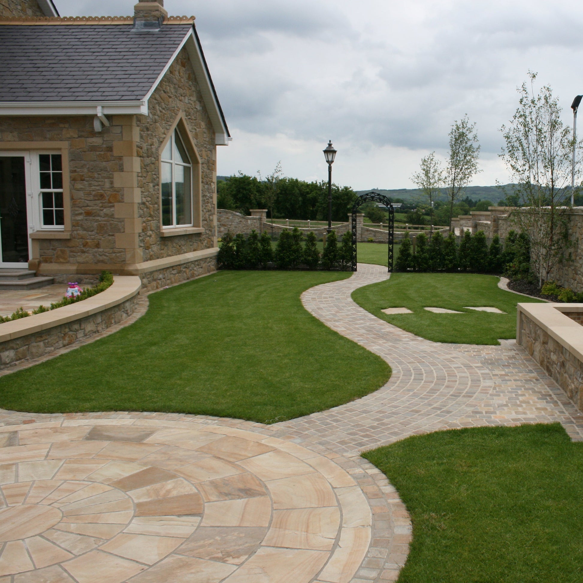 Tinted Mint Sandstone Circle with cobbled pathways on a well maintained garden with hedging and planting outside a large stone house. 